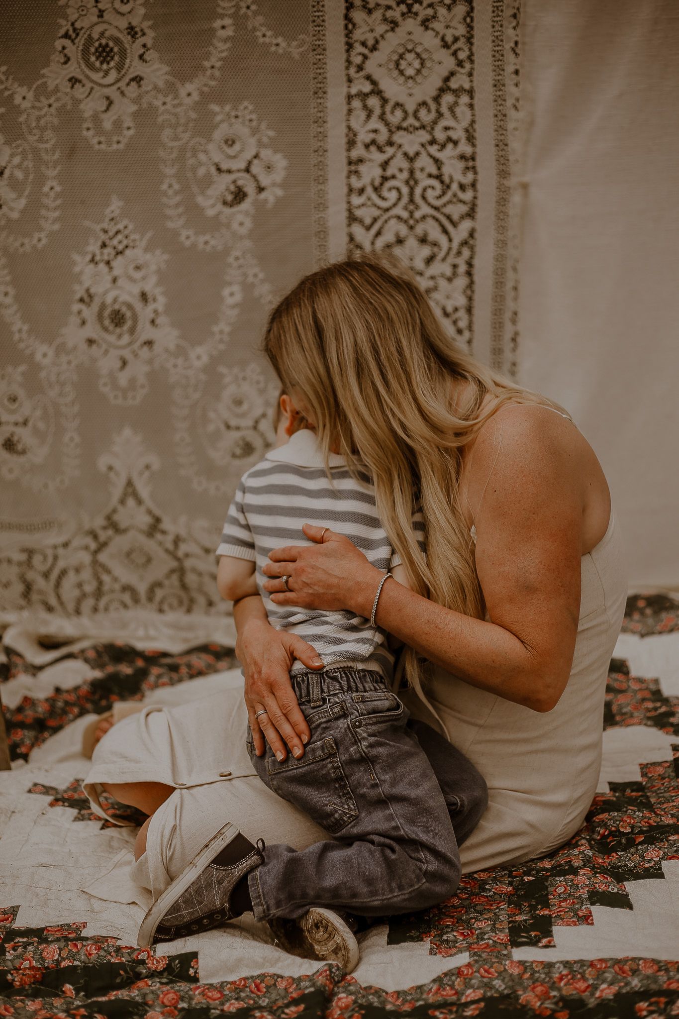 A mother with long blonde hair sits on a quilt hugging her young child in a striped shirt before a decorative tapestry.