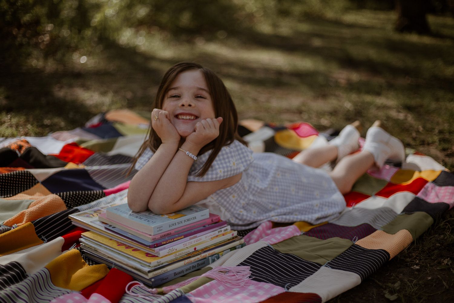 Young girl smiling while lying on a colorful patchwork quilt outdoors, resting her chin on a stack of books.