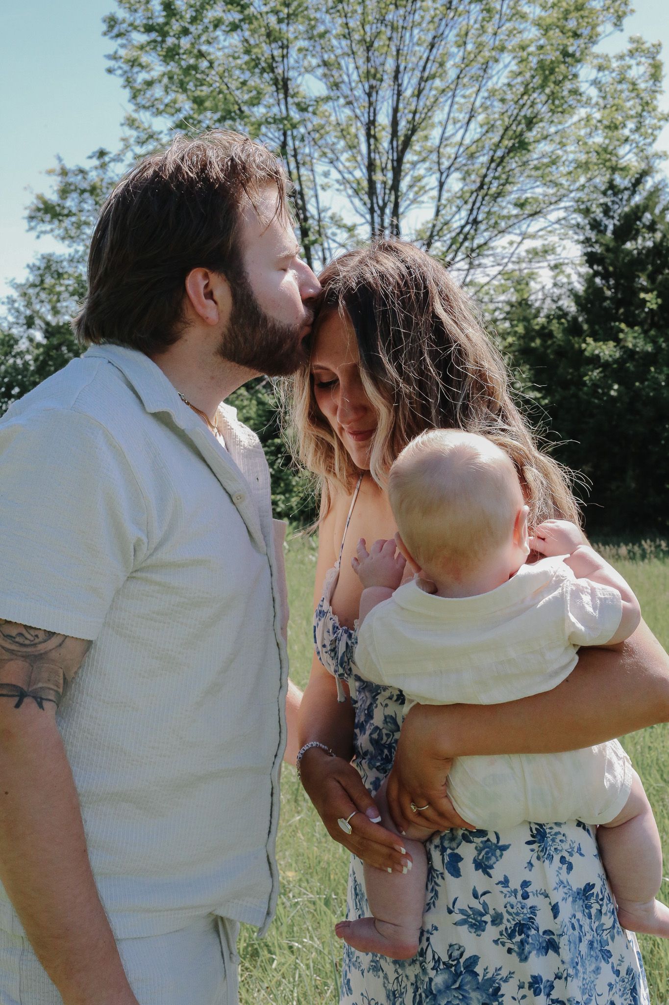 A bearded man kisses a woman's forehead as she holds their baby in a sunny green field.