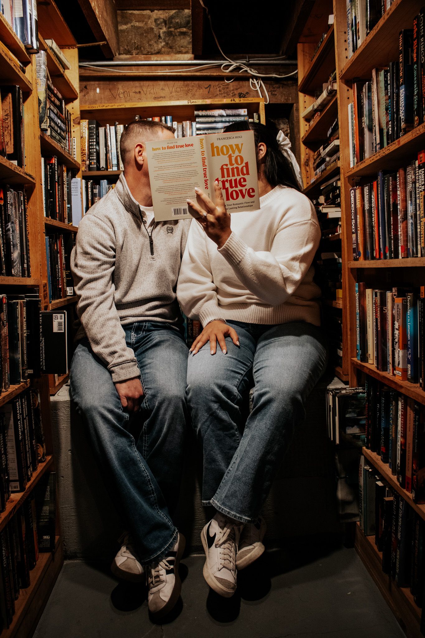 Couple sitting in a cozy bookstore aisle, faces hidden behind a book titled How to Find True Love.