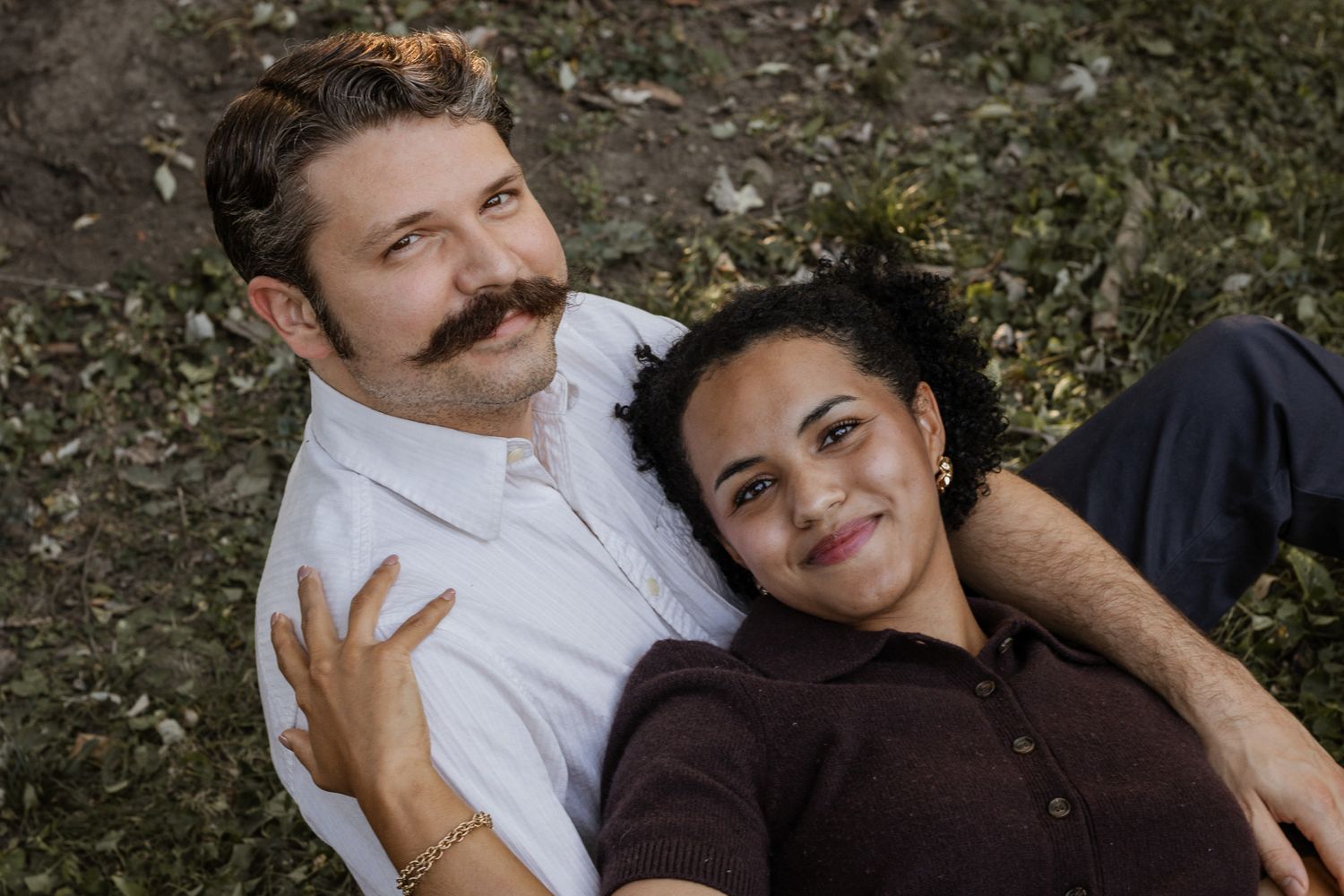 A smiling couple relaxes together on grass, the woman resting her head on the mans shoulder.