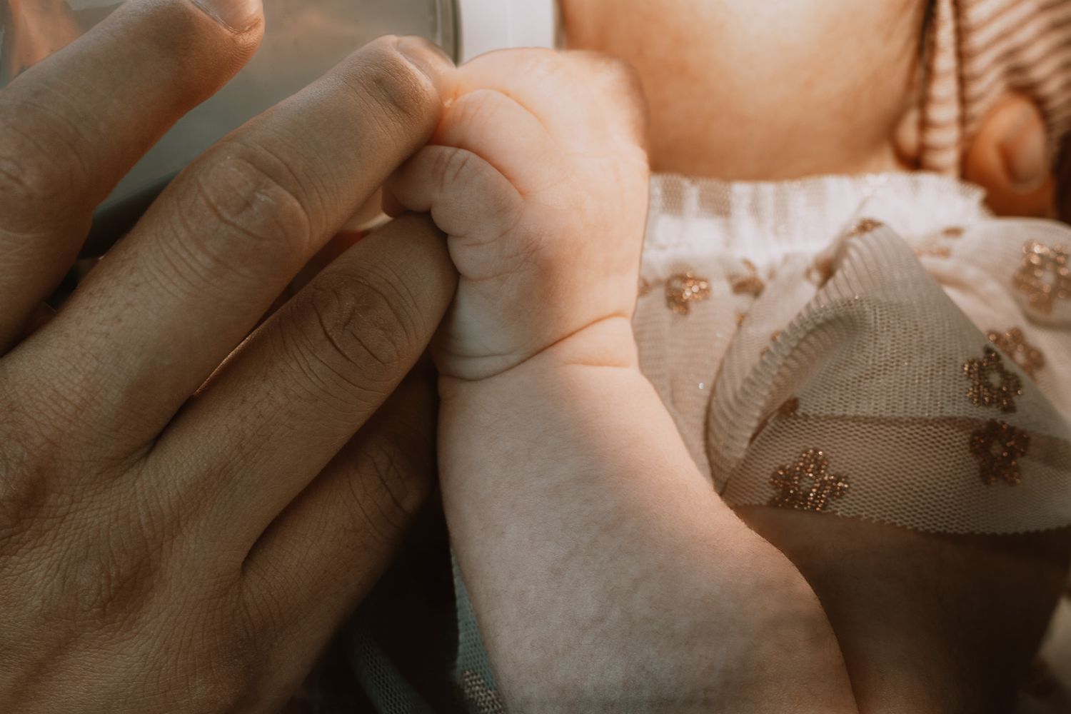 Close-up of newborn baby gently grasping adult finger wearing delicate floral embroidered outfit.