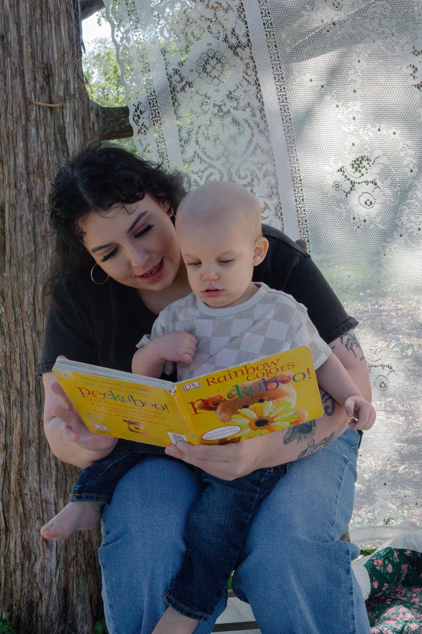 Woman with dark hair reads a yellow Peekaboo book with a bald baby on her lap outdoors.