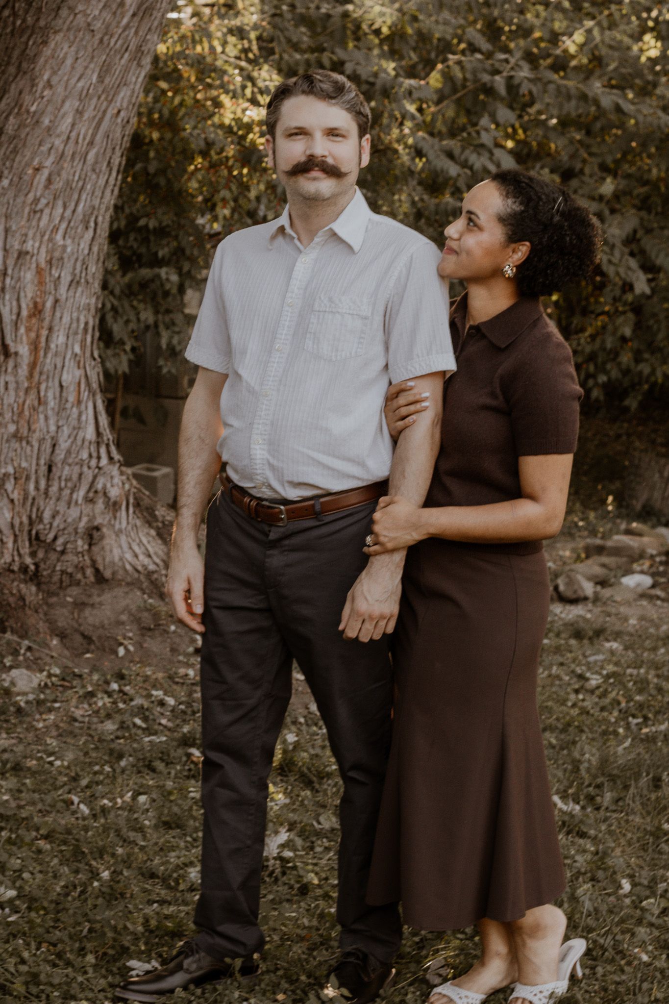 A couple poses outdoors near a tree, man in white shirt with mustache, woman in brown dress smiling up at him.
