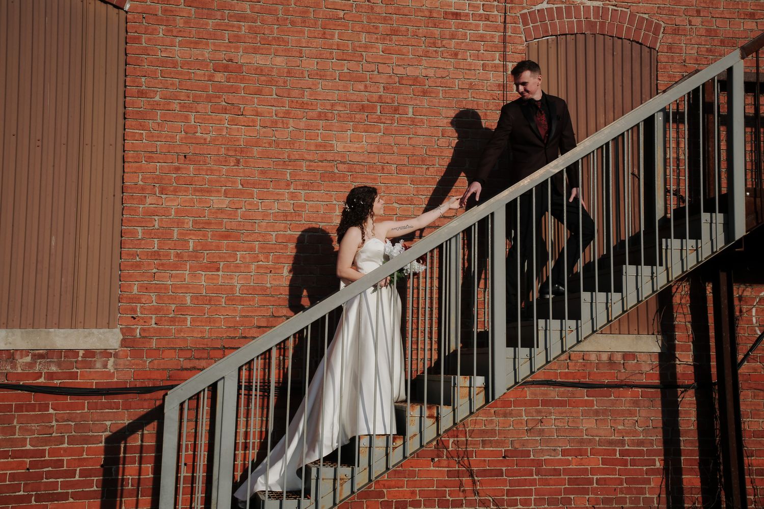Bride and groom holding hands on outdoor metal staircase against red brick wall in wedding attire.