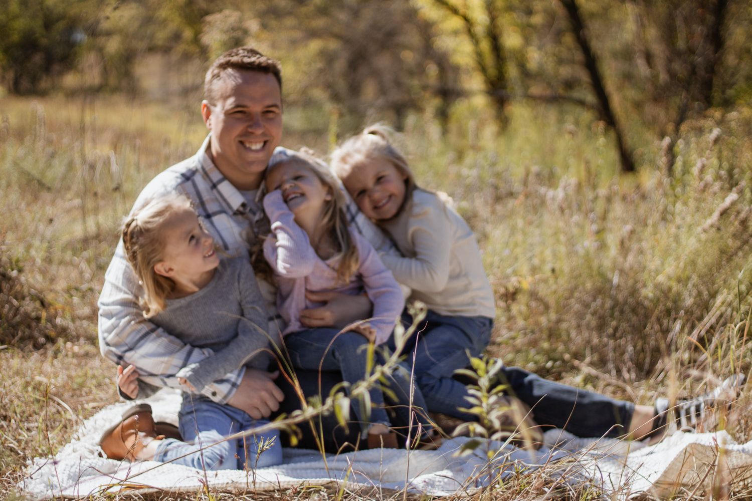 Father sitting in an autumn field smiling with his three daughters hugging him during a fall family photo session.