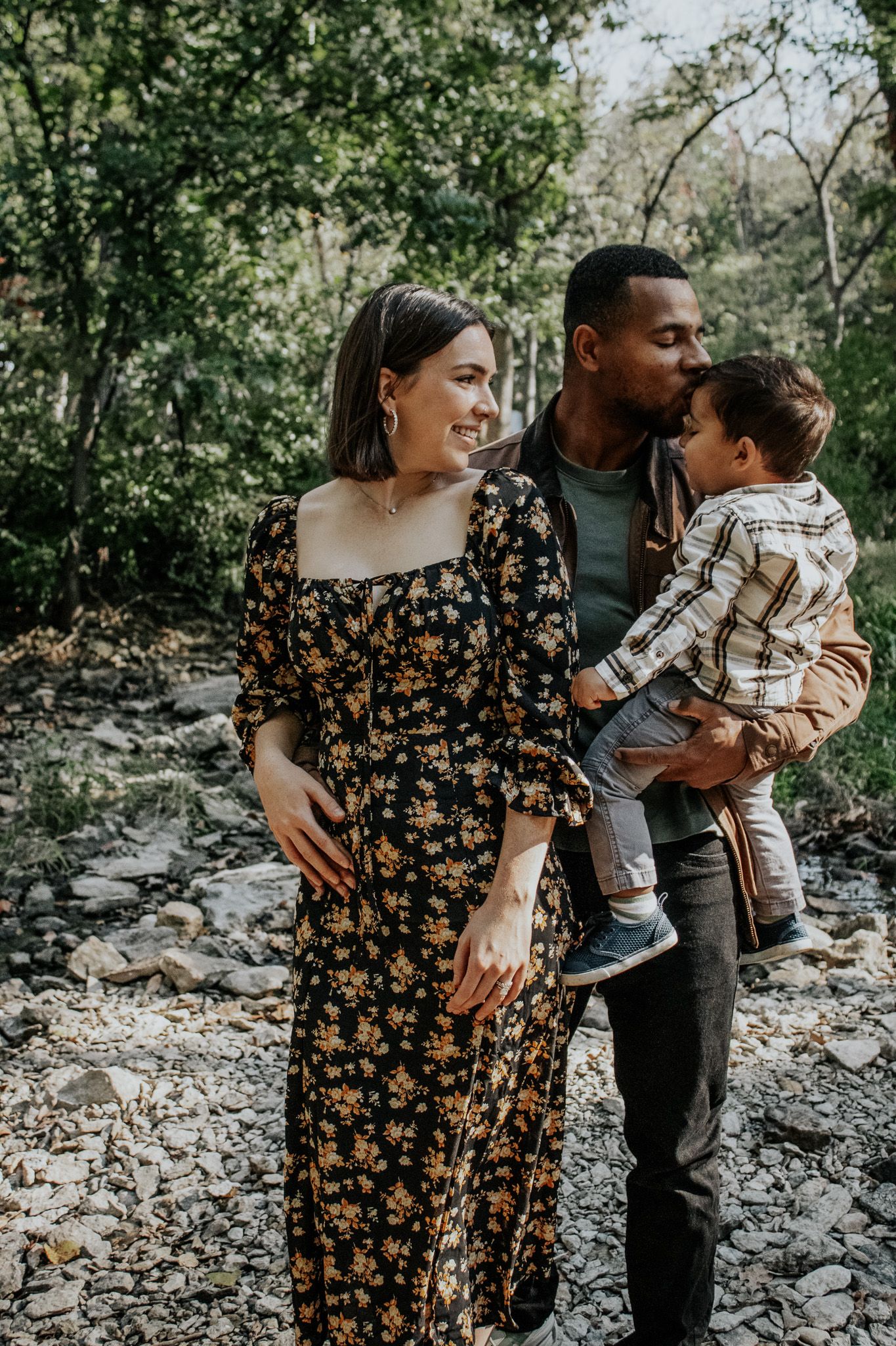 Interracial family with a toddler boy posing together on a rocky nature trail surrounded by green trees.