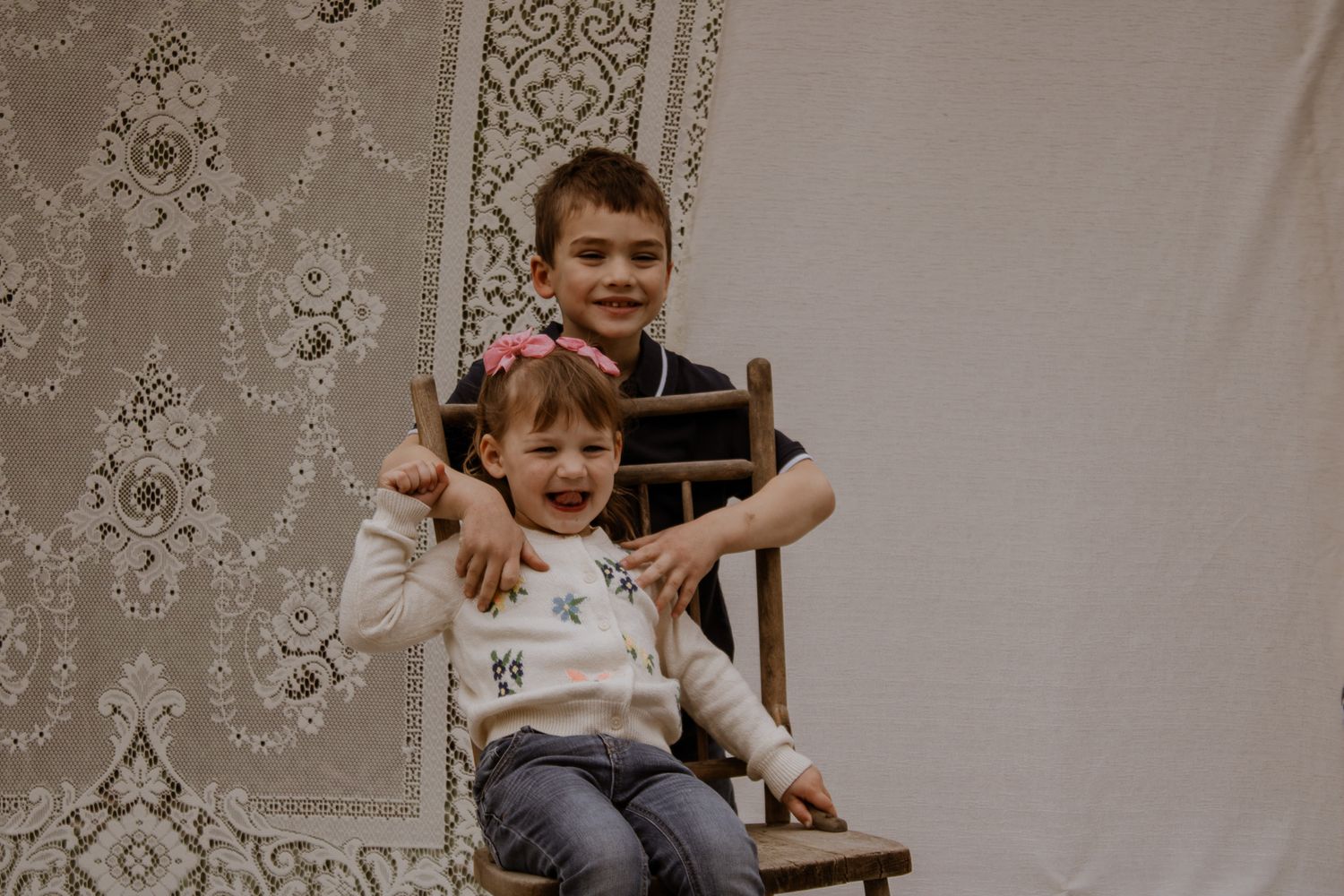 Two happy children posing together on a wooden chair in front of a decorative backdrop.