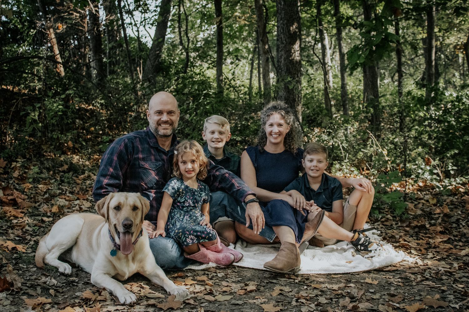 Happy family of five with a yellow Labrador dog sitting together on fallen leaves in an autumn forest setting.