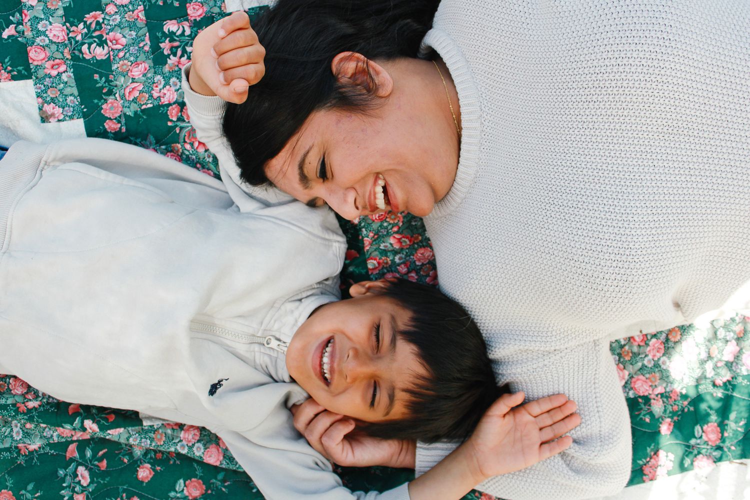 Two laughing children and a woman lying on a floral blanket outdoors, sharing a joyful moment together.