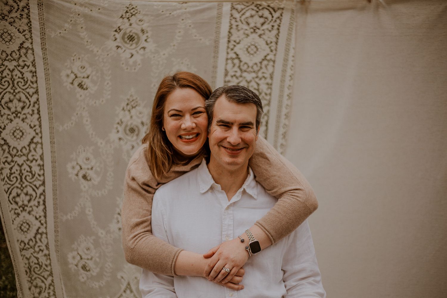 A smiling couple posing together, woman hugging man from behind, in front of a decorative lace backdrop.