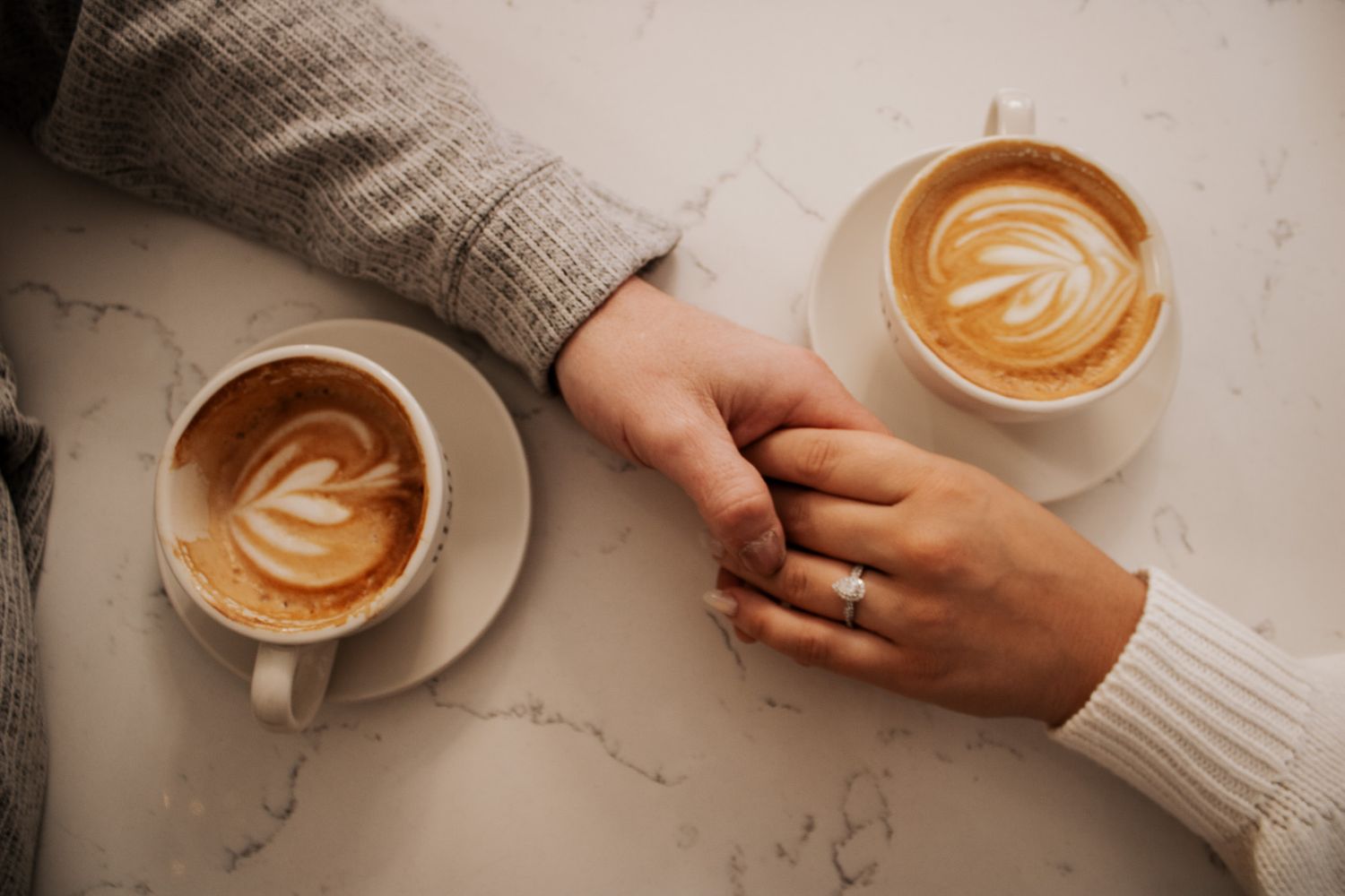 Couple holding hands over a marble table with two lattes, one wearing a diamond engagement ring.