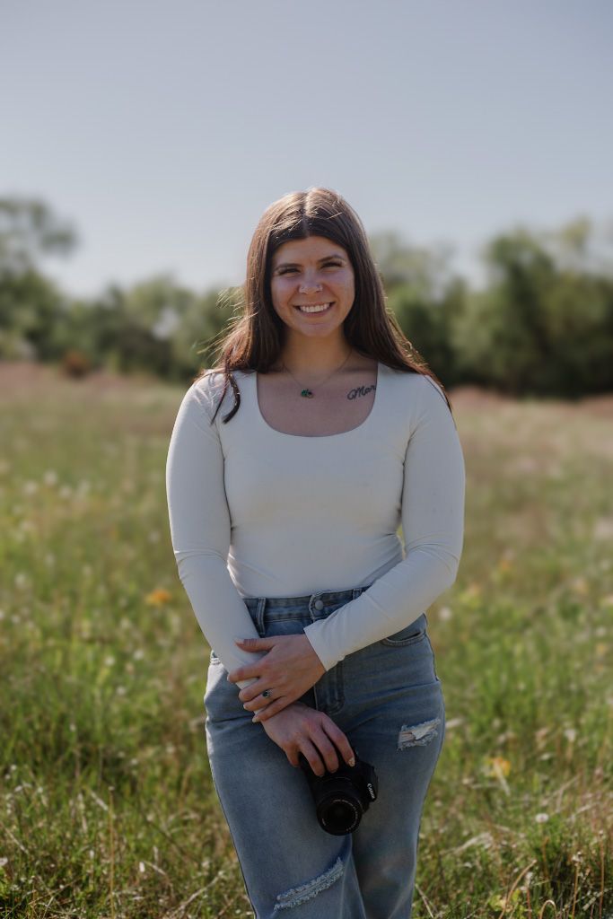 Young woman smiling in a field, wearing a white long-sleeve top and jeans, holding a camera outdoors.