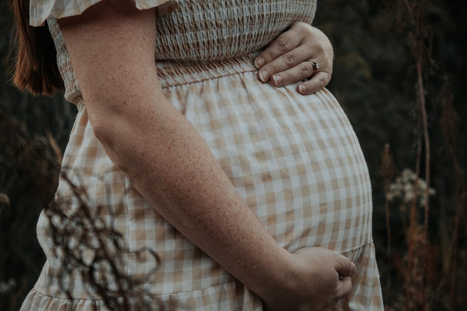 Close-up of a pregnant woman in a checkered dress cradling her baby bump outdoors.