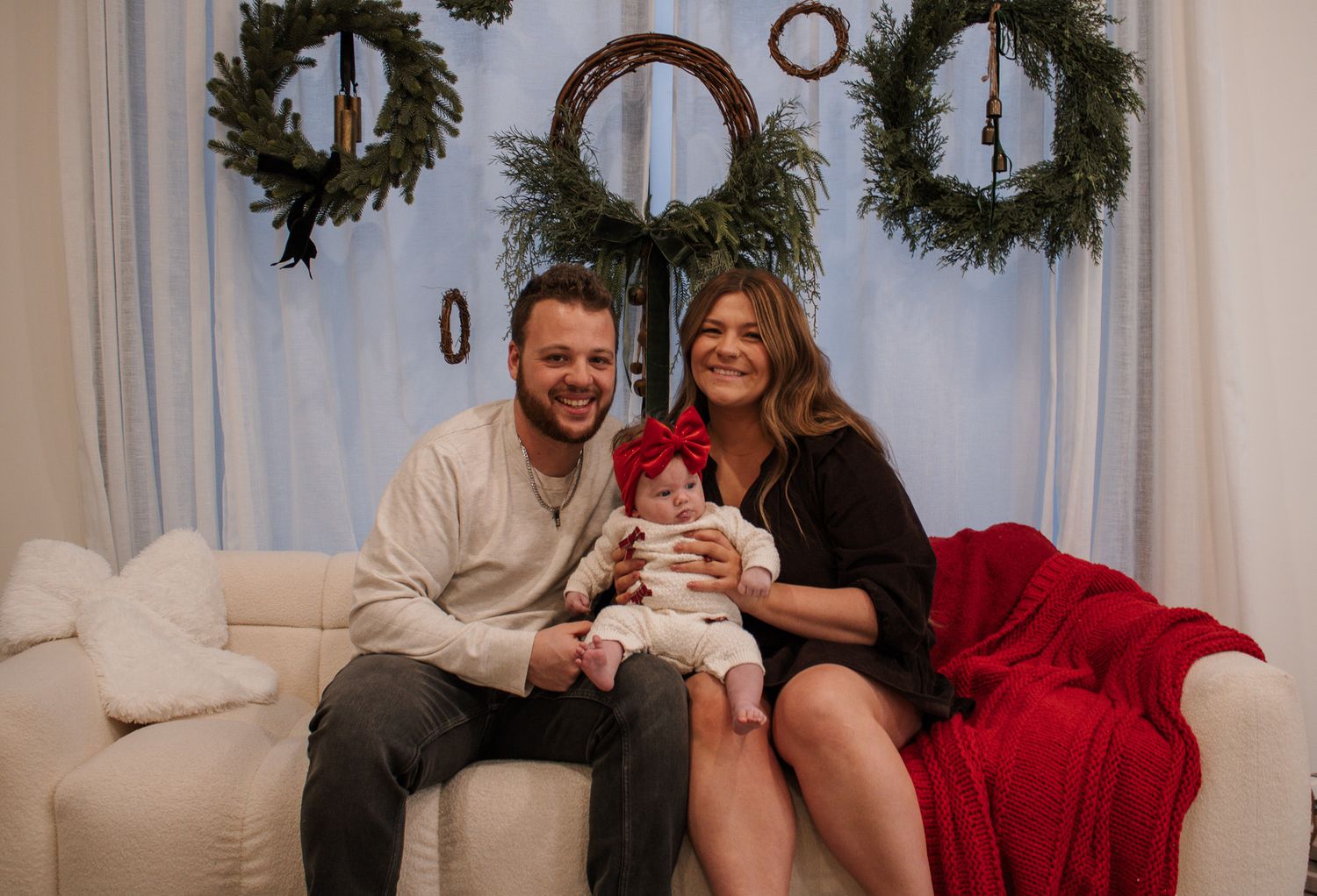 A smiling couple sits on a white couch holding their baby in a red bow during a festive Christmas photo session.
