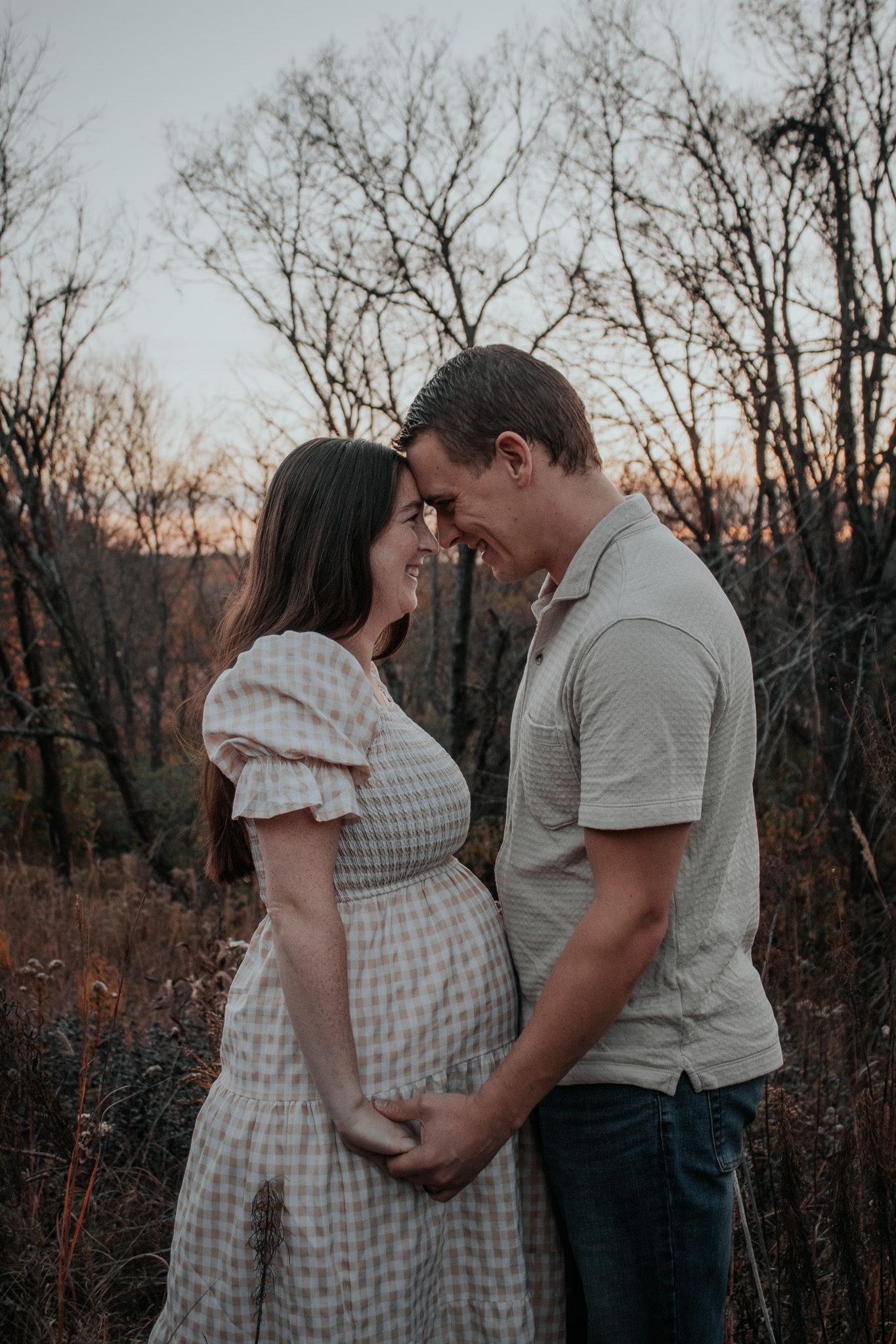 Expecting couple holding hands and touching foreheads at golden hour in a natural setting.