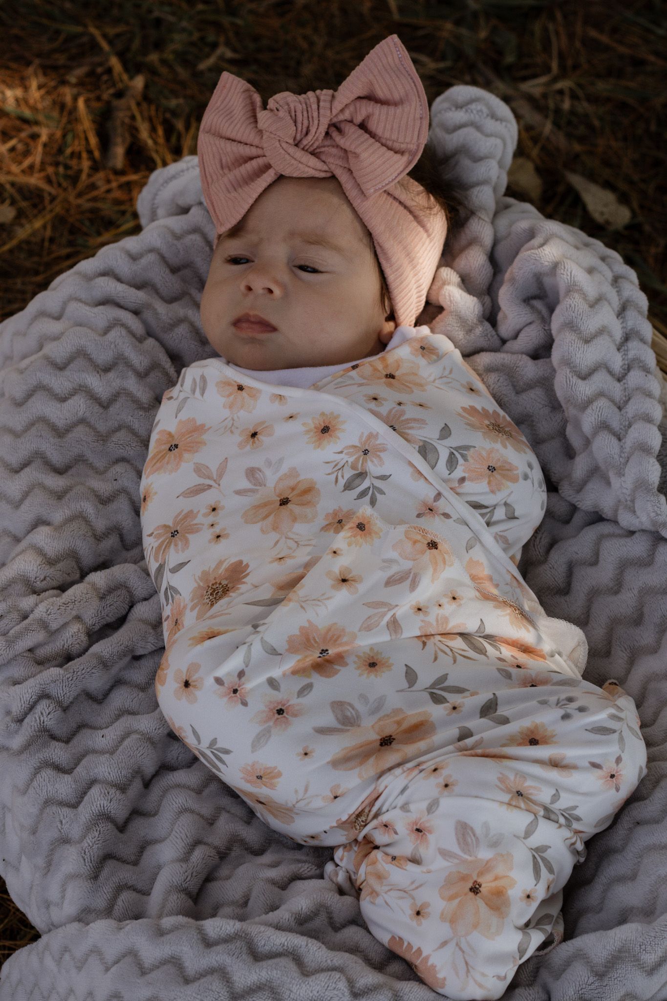 Sleeping newborn baby swaddled in a floral blanket wearing a pink bow headband outdoors.