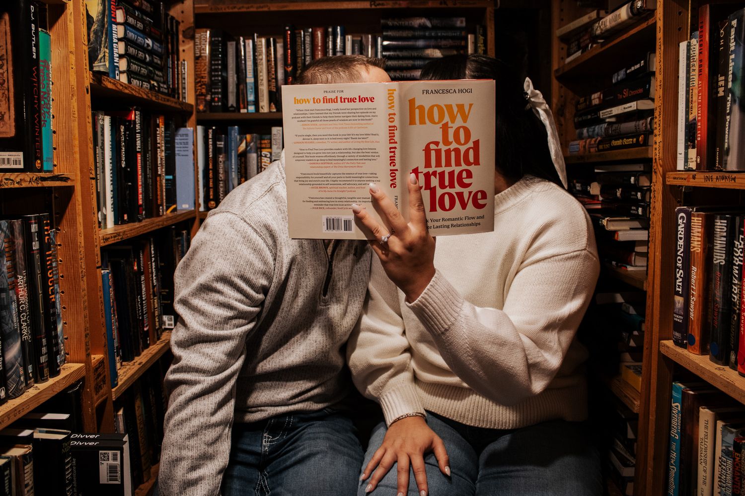 Couple hiding faces behind 'How to Find True Love' book while sitting together in a cozy bookstore.
