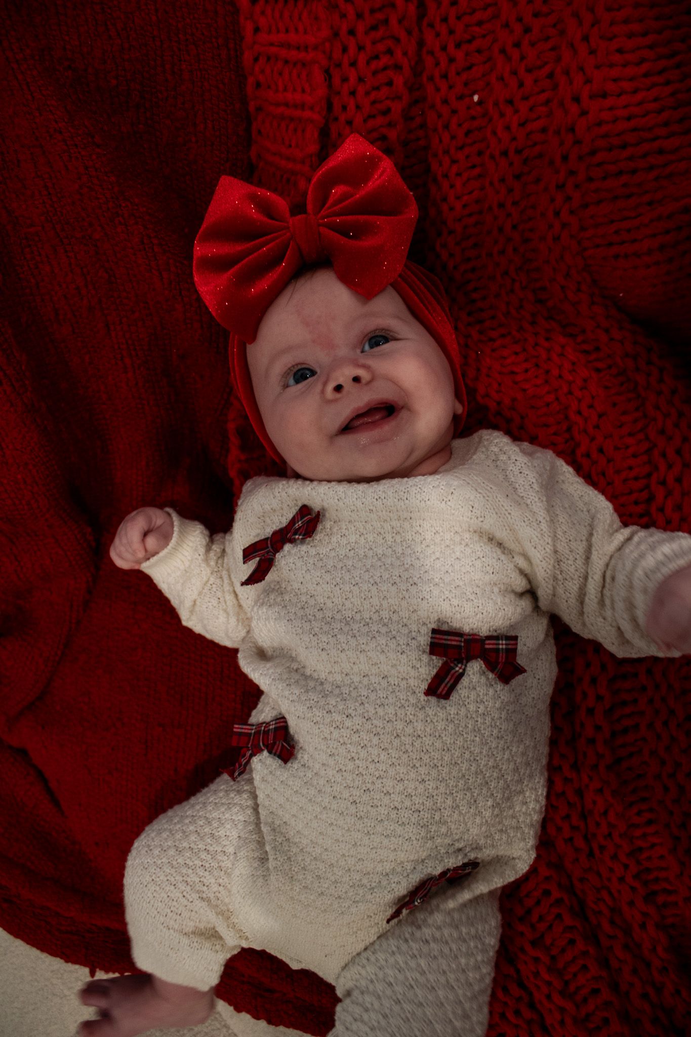 Smiling newborn baby wearing a cream knit outfit with red bows and a large red bow headband.