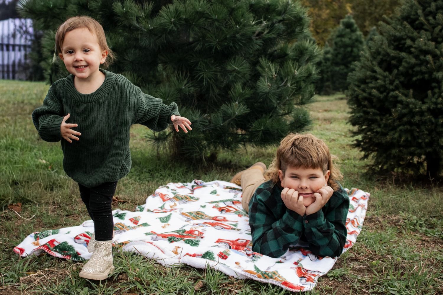 Two young children playing outdoors near Christmas trees, one lying on a holiday blanket and one standing and smiling.
