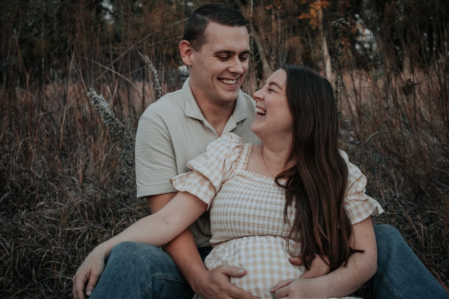 Smiling couple sitting in a field, pregnant woman cradling her baby bump during an outdoor maternity photo session.