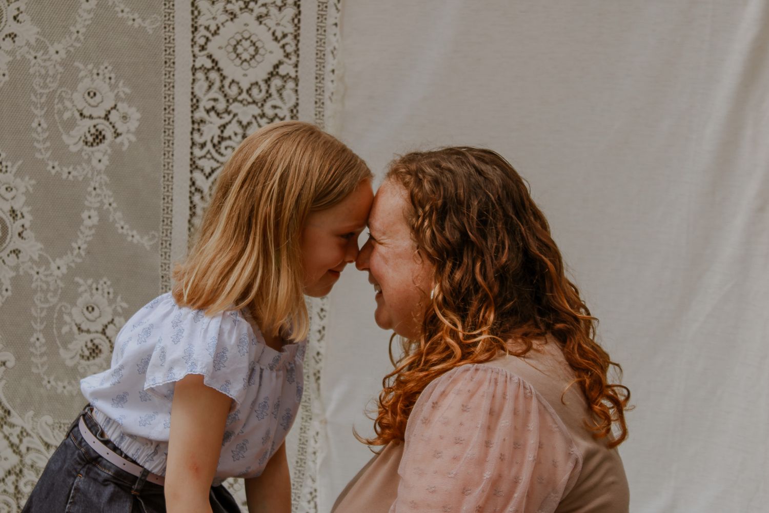A mother and young daughter touch foreheads lovingly in front of a lace backdrop.