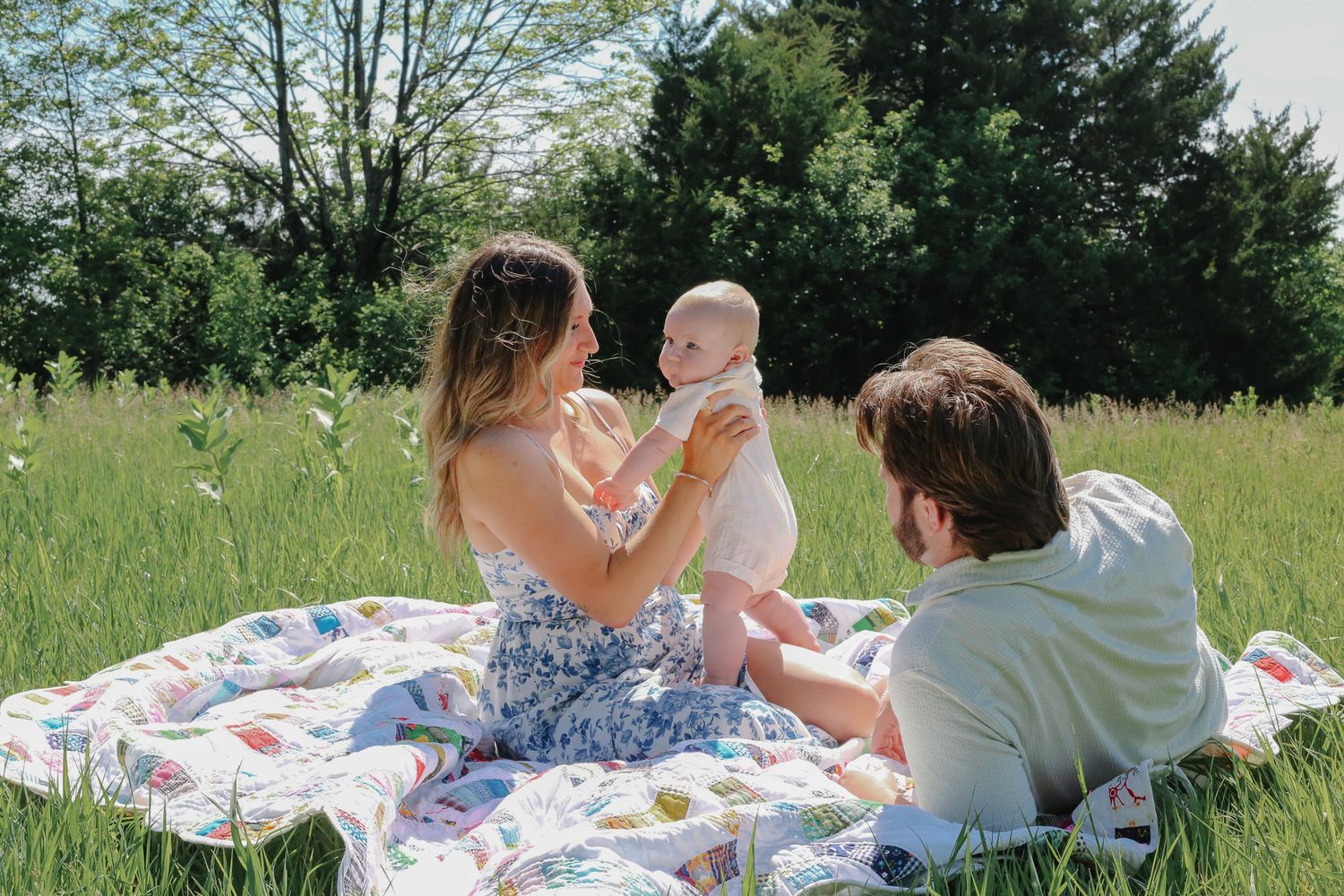 A young family enjoys a sunny picnic in a green meadow, with parents holding their baby on a colorful quilt.