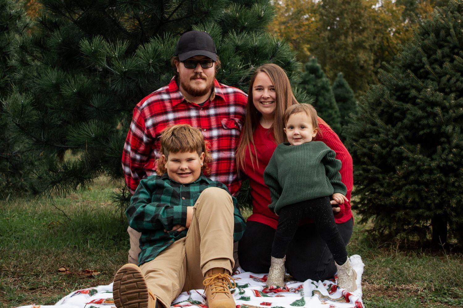 A happy family of four sits together on a Christmas blanket at a tree farm.