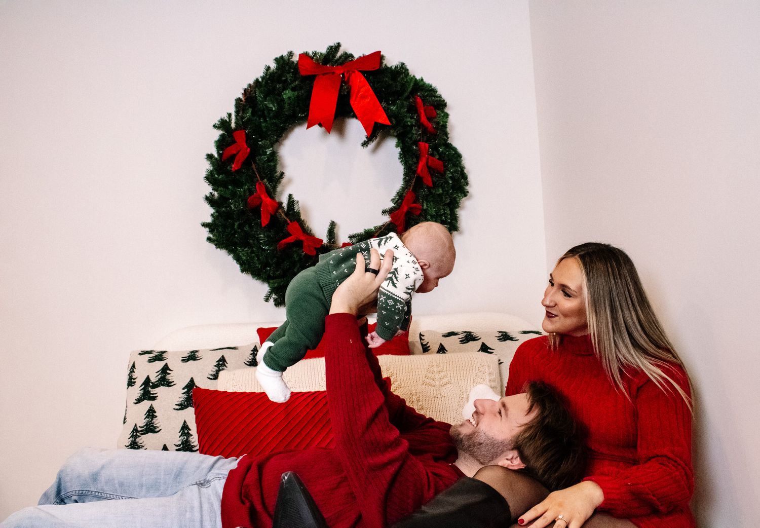 Family celebrating Christmas with parents lifting baby in festive holiday outfits near wreath.