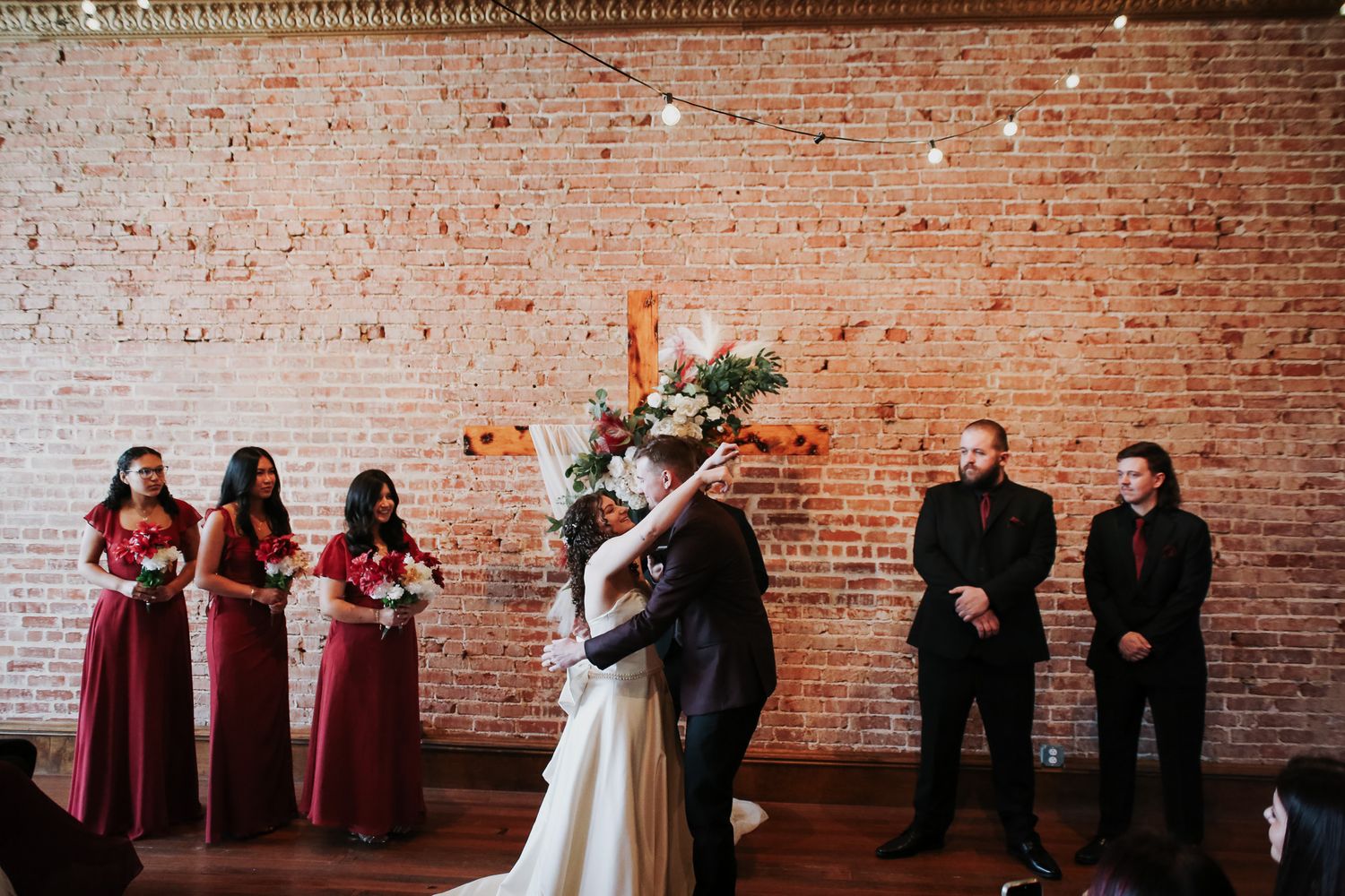 Bride and groom share first kiss at rustic brick wall wedding ceremony with bridesmaids in red and groomsmen nearby.