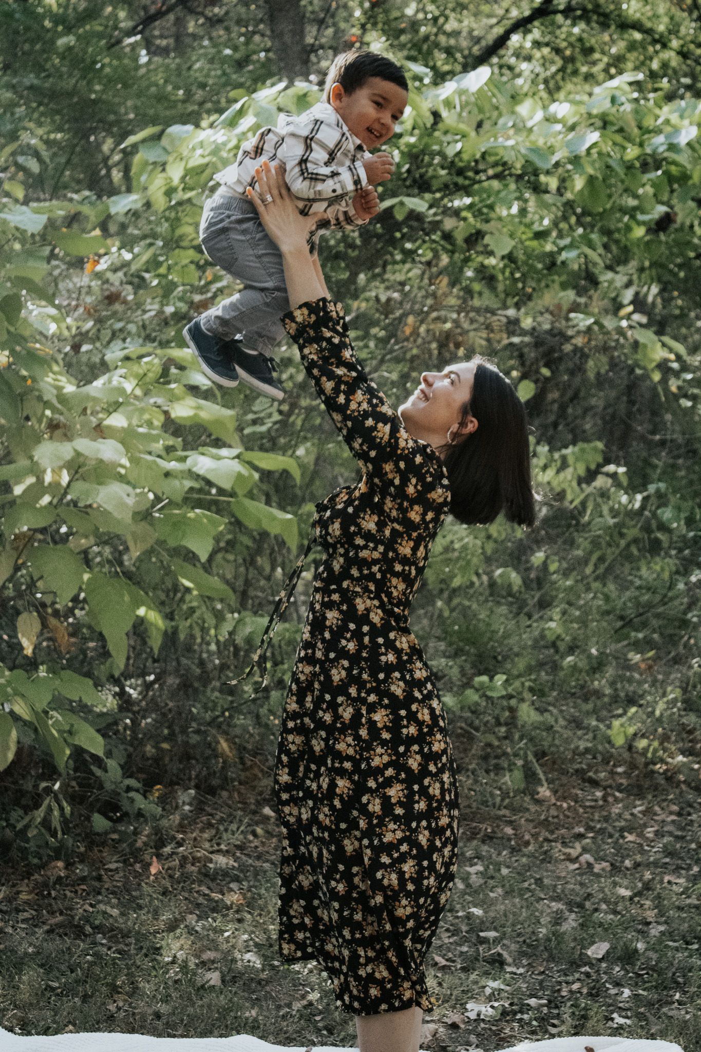 Happy mother lifting her toddler son high in the air surrounded by lush green trees.