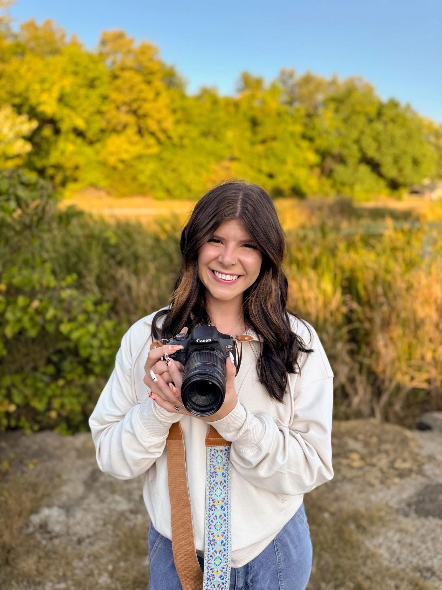 Smiling brunette photographer holding a Canon camera outdoors with green trees in background.