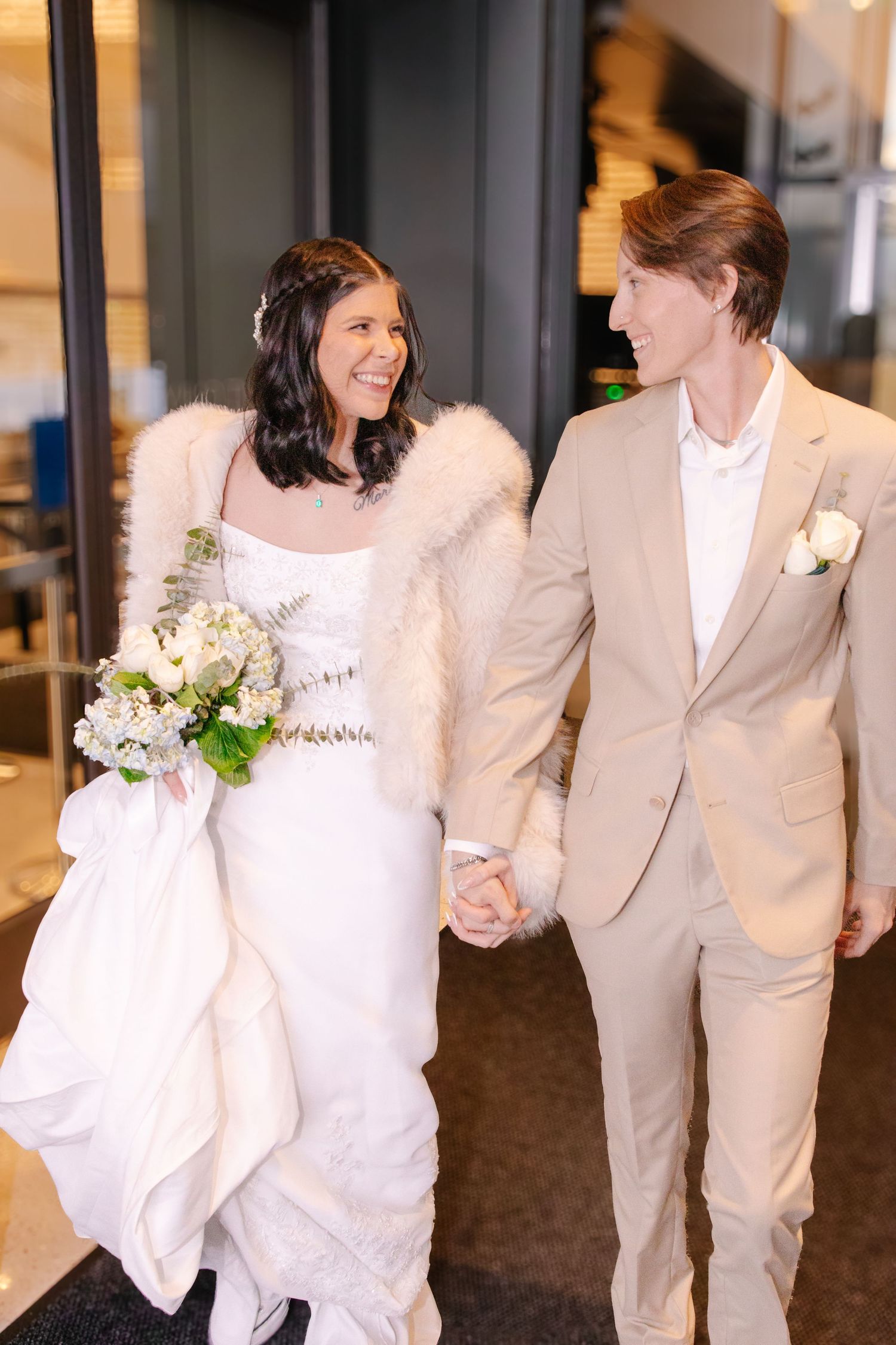 Happy bride and groom walking hand in hand, bride wearing fur stole and holding white bouquet.