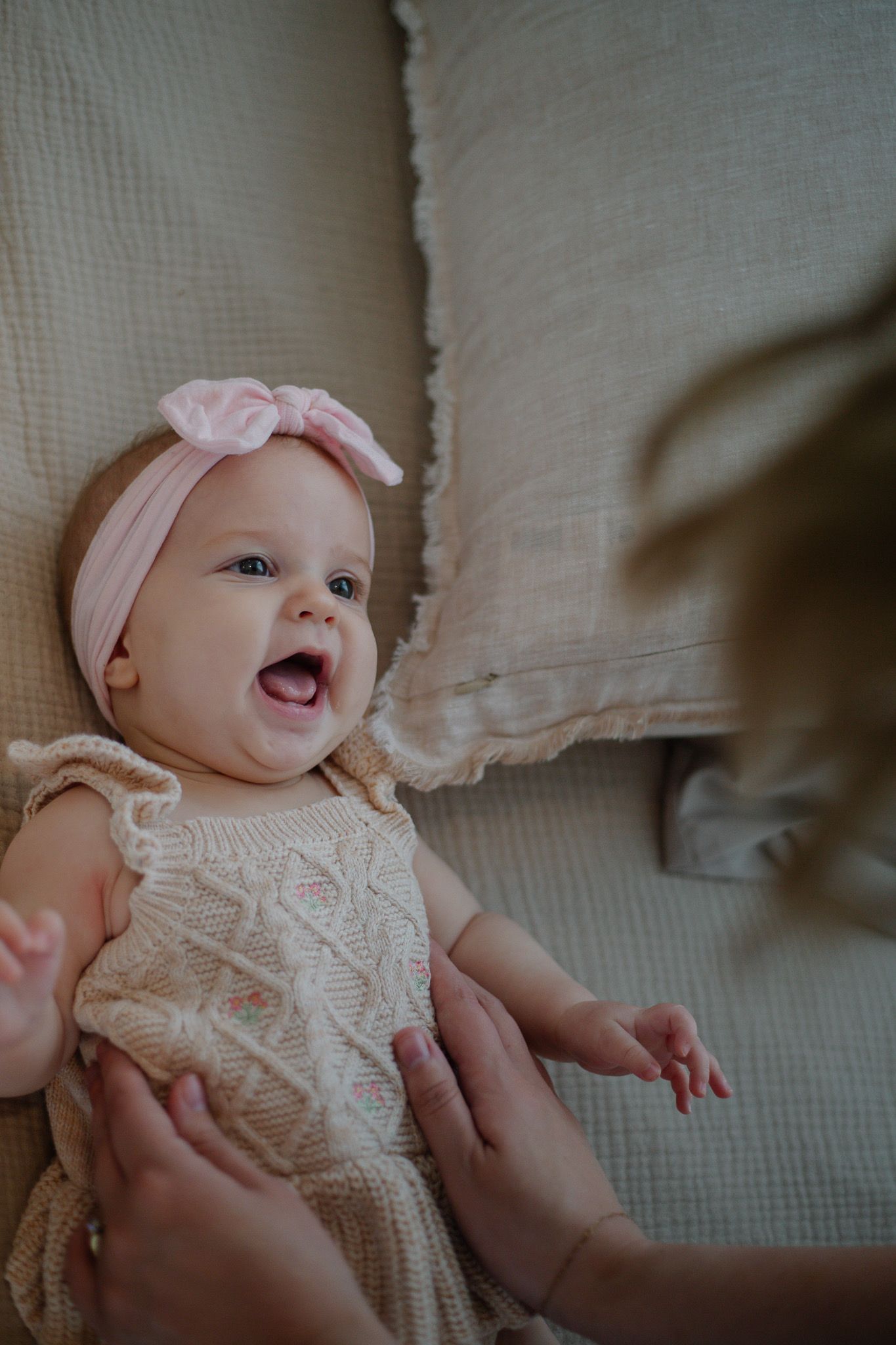 Happy baby girl in pink bow headband and knit dress laughing while lying on a couch cushion.