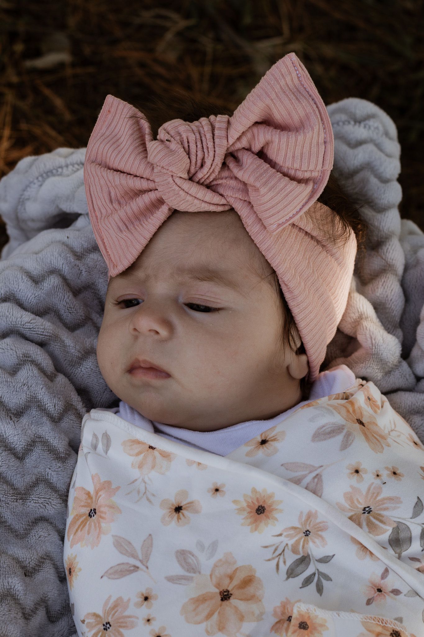 Newborn baby wrapped in floral swaddle wearing pink bow headband rests on gray chevron blanket.