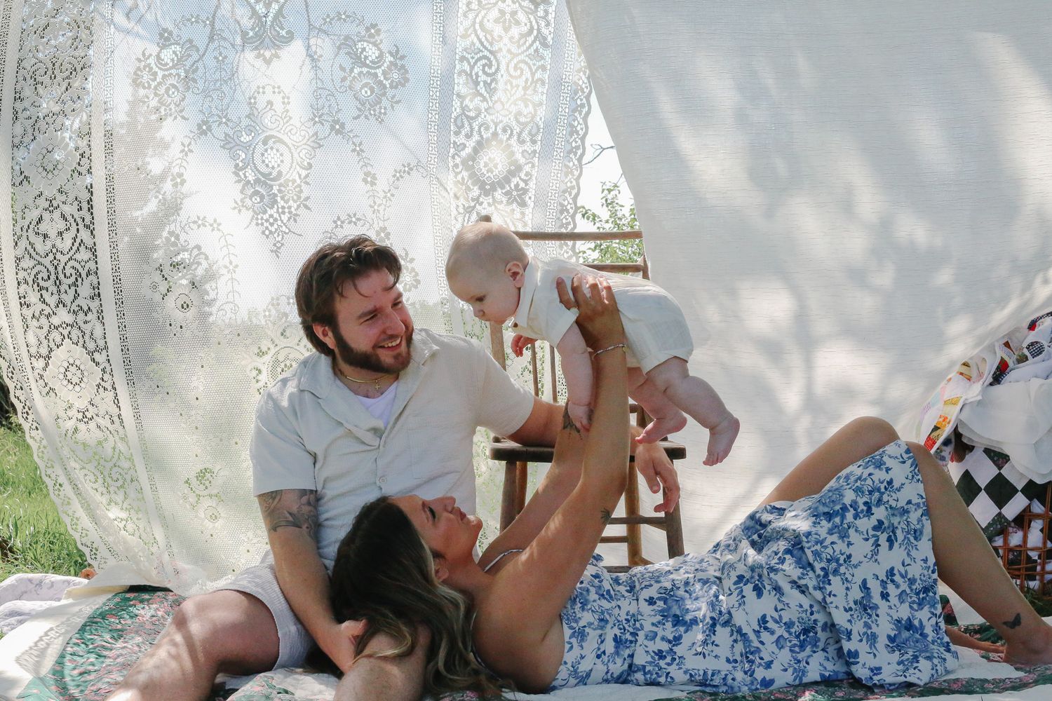 A couple relaxes outdoors under white lace curtains, with the man sitting and woman lying down lifting their baby in the air.