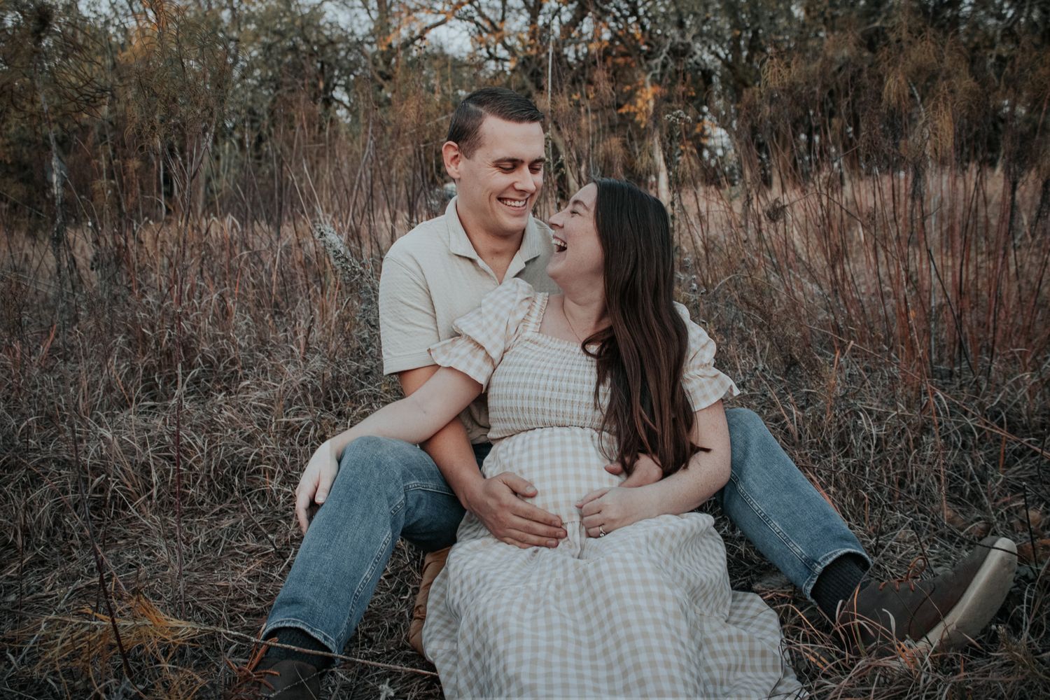 Happy expecting couple sitting together in a field, smiling and cradling the baby bump.