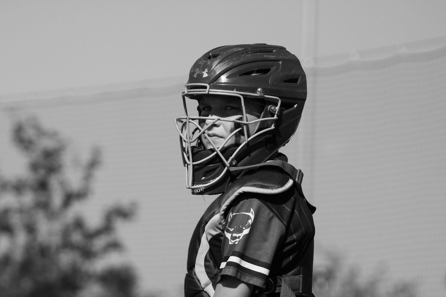 Black and white photo of a youth baseball catcher in full gear, including helmet and chest protector, standing in profile.