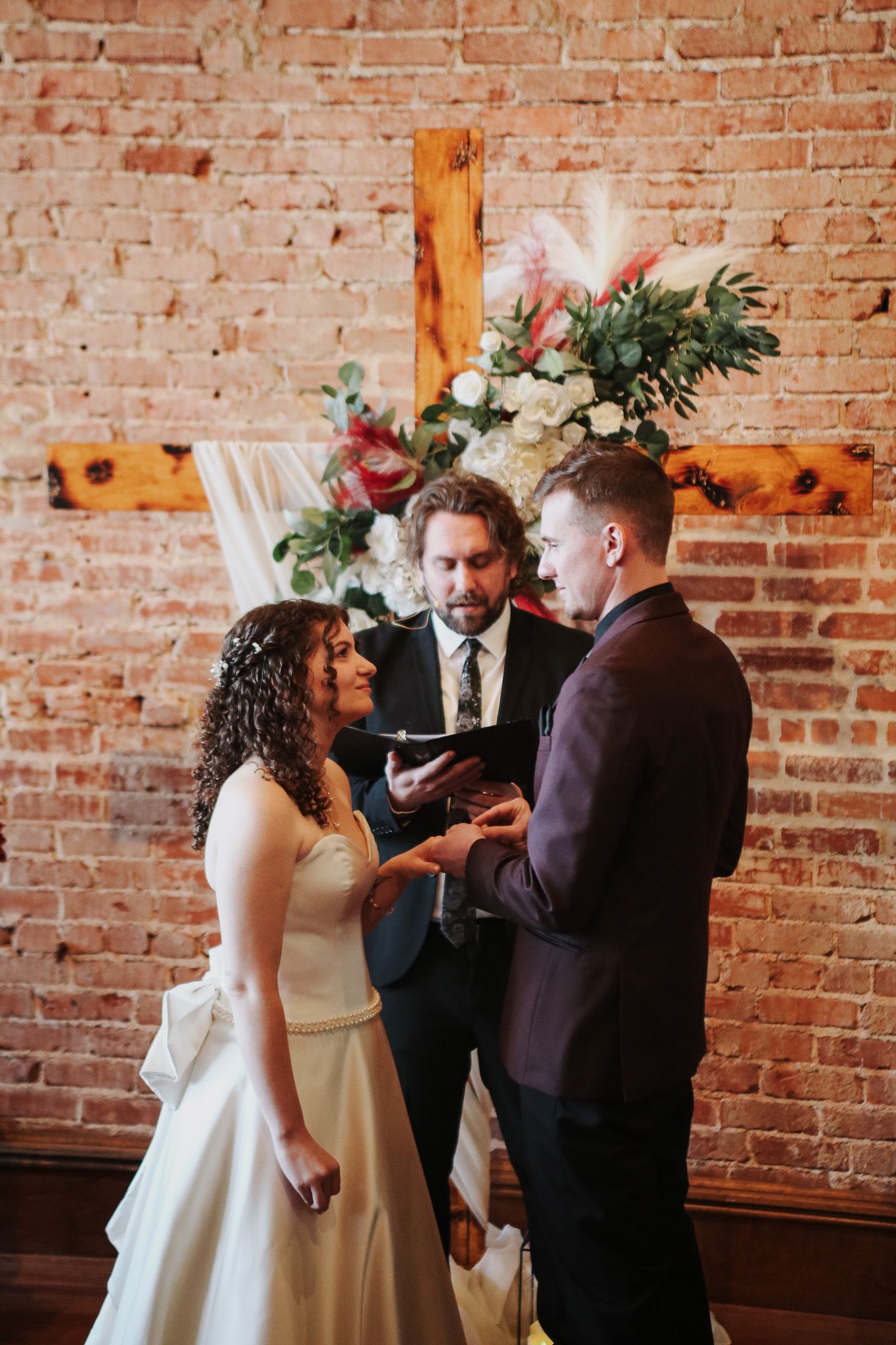 Bride and groom exchange rings during indoor wedding ceremony before brick wall with wooden cross and floral arrangement.