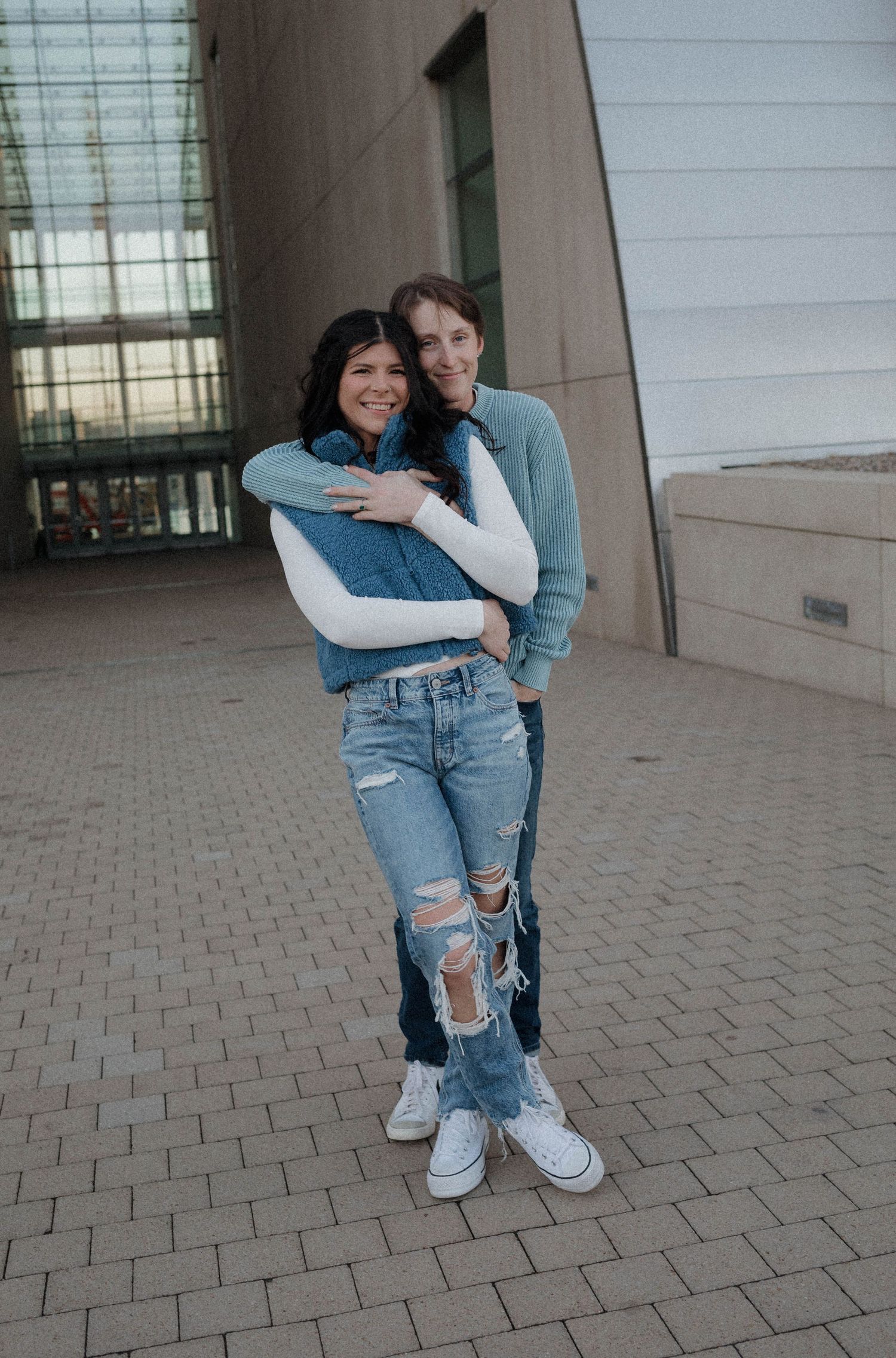 Couple posing together outdoors, one embracing the other from behind near modern building.