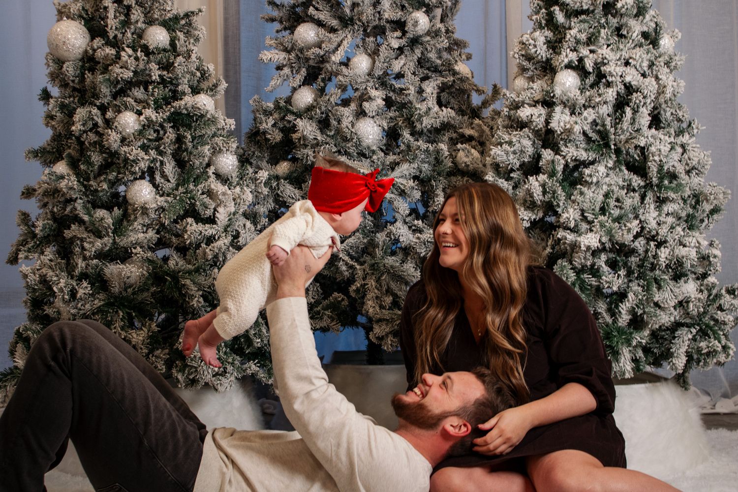 Couple sitting on floor lifting baby in Santa outfit in front of snowy Christmas trees holiday photo.