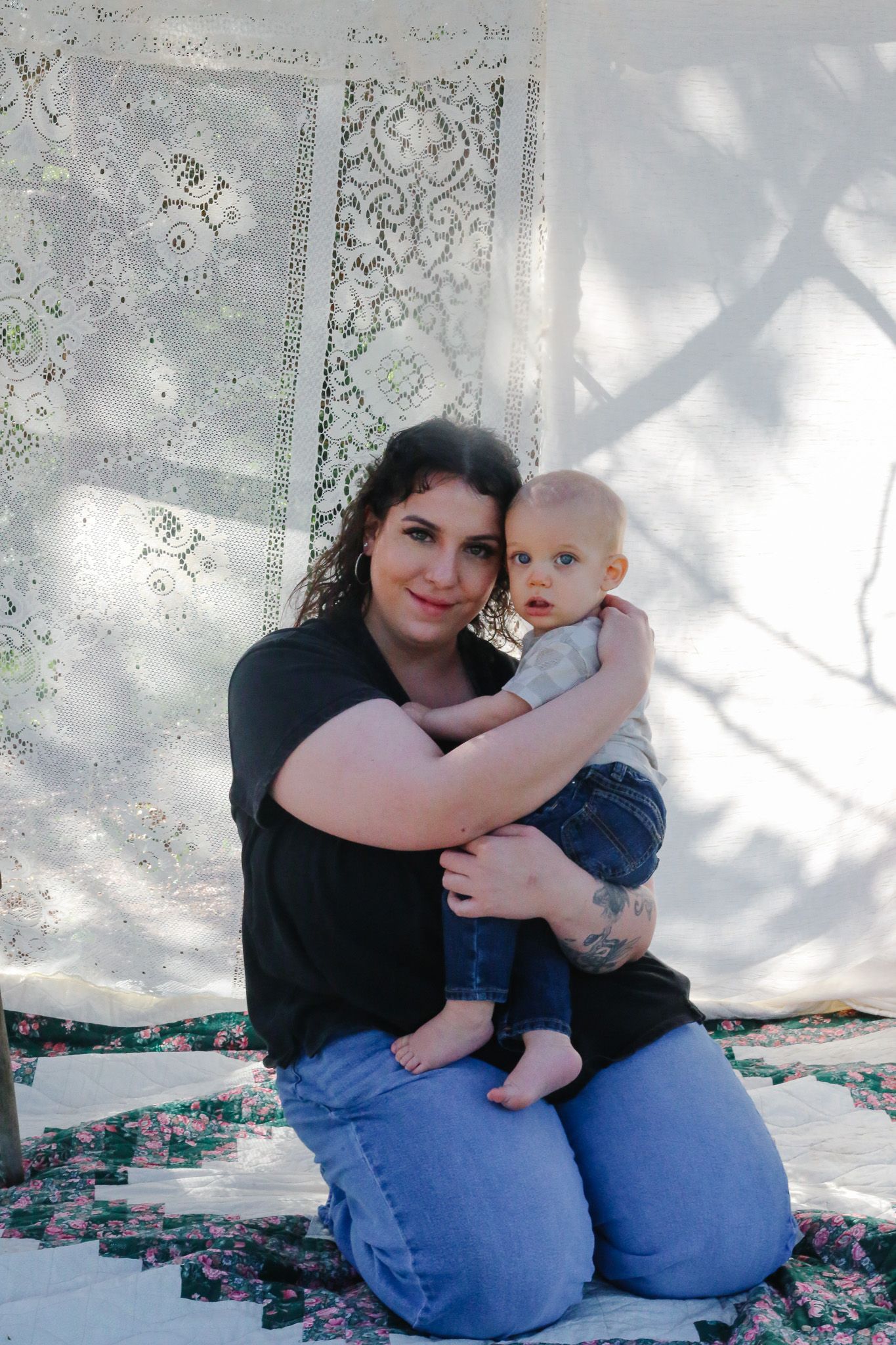 A young woman in a black shirt and jeans kneels on a quilt, lovingly holding a baby in front of a lace backdrop.