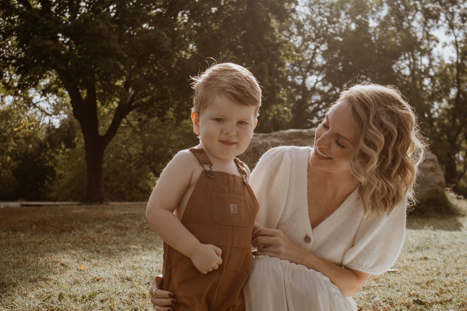 A mother in a white dress holds her toddler boy in brown overalls outdoors in a sunlit park.