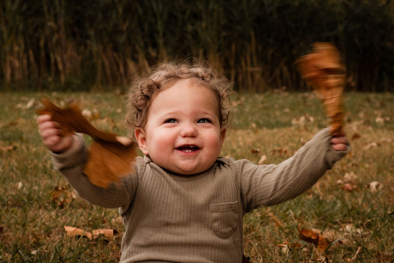 A small child in a beige sweater plays joyfully outdoors among fallen autumn leaves.