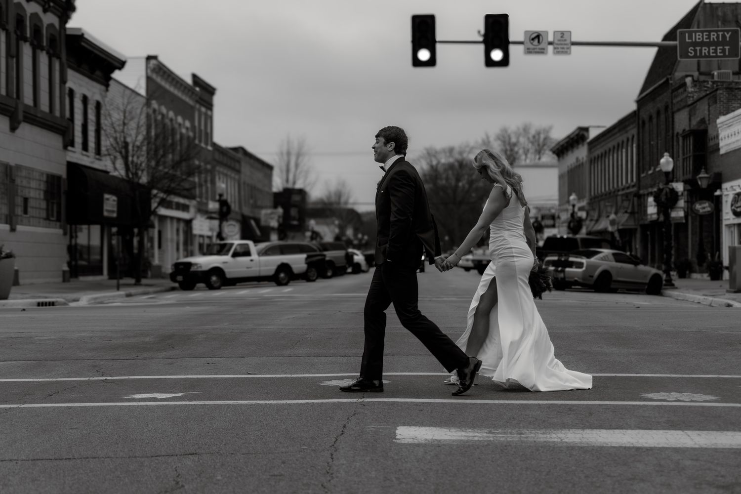 A couple in wedding attire crosses a small town intersection in black and white.