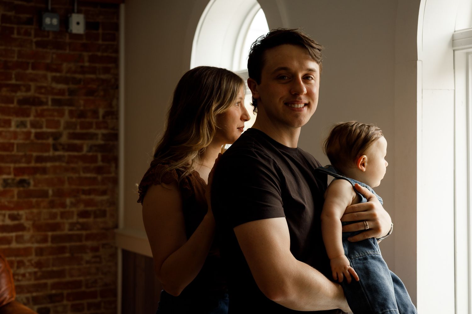 A young family poses indoors, with a man holding a baby and a woman leaning against him near a brick wall.
