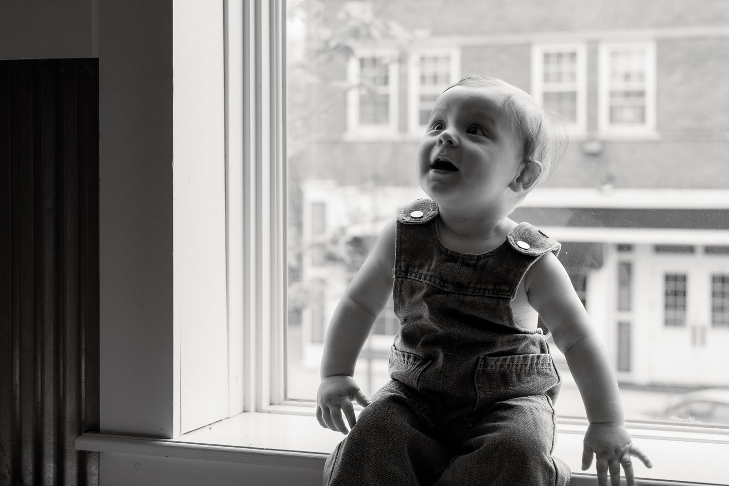 Happy baby sitting on windowsill laughing in black and white portrait photo.
