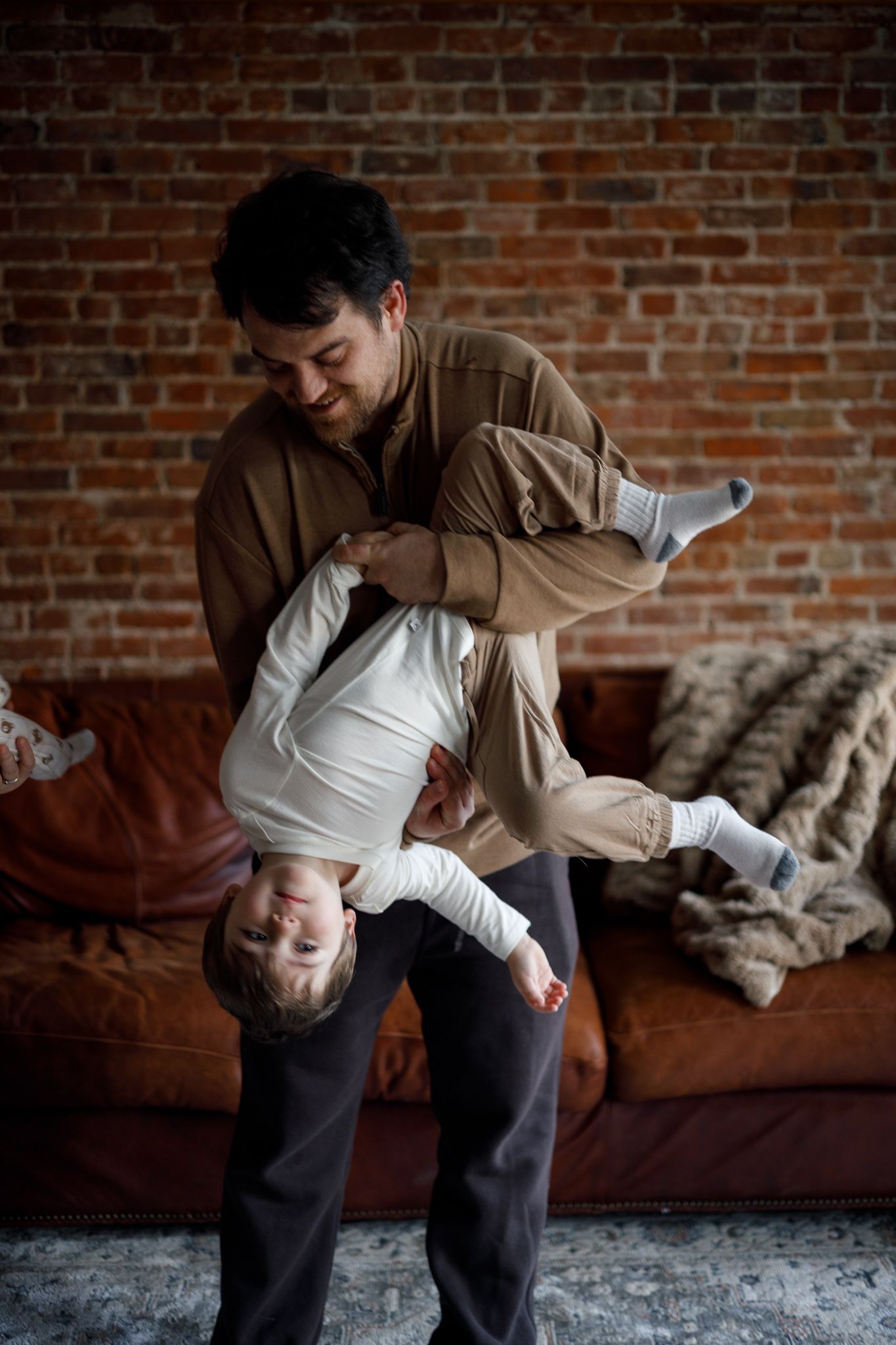 Two people share a playful moment dancing and dipping on a brown leather couch against an exposed brick wall.