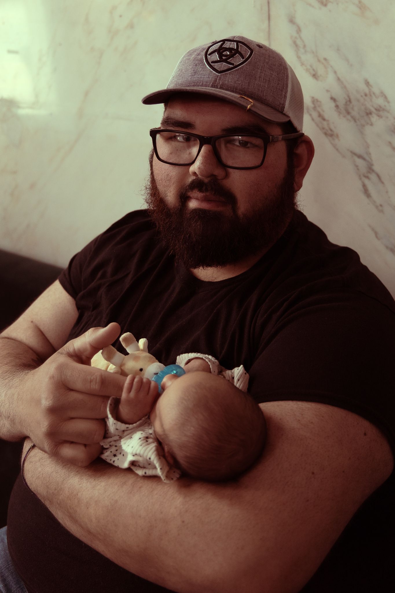 A bearded man in glasses and a baseball cap holds a sleeping newborn baby in a series of tender moments.