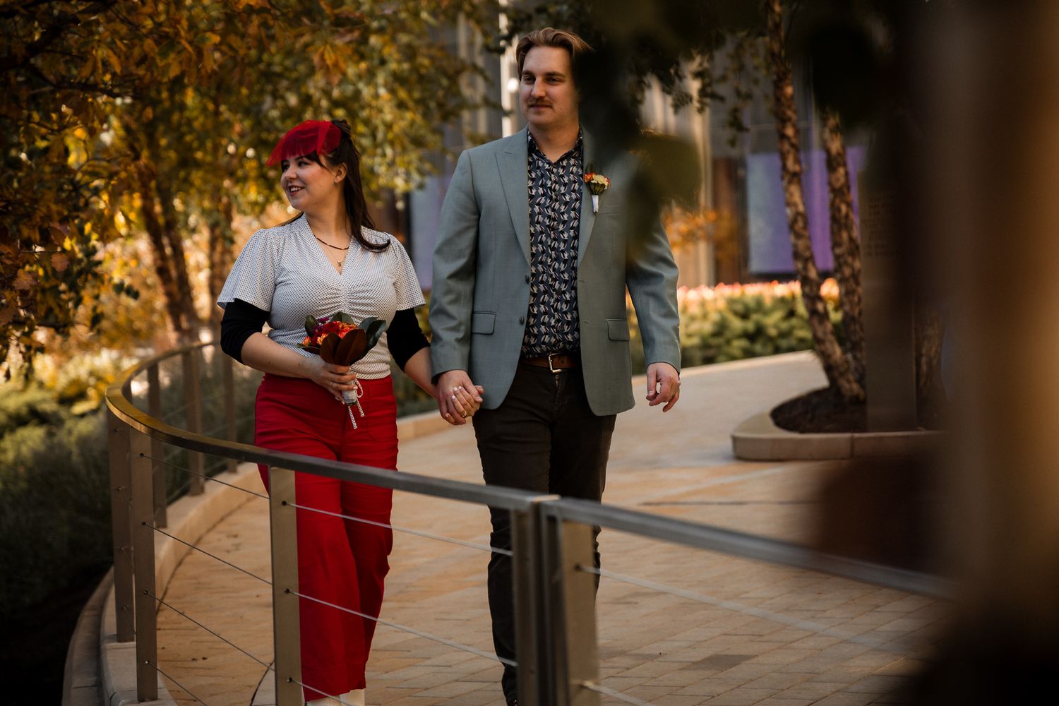 A couple in elegant attire walks together through an autumn garden setting during a photo session.